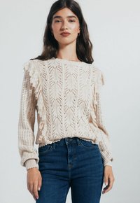 Donna che indossa un maglione a maglia color crema con volant sulle maniche e jeans blu a vita alta, in piedi di fronte a uno sfondo neutro.