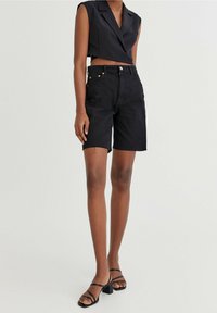 Shorts in denim nero con vita alta, orlo sfilacciato e cinque tasche. Abbinati a un top corto senza maniche nero e tacchi neri con cinturini.