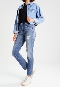 Svetle modrá riflová bunda s černým tričkem, doplněná o světlé modré džínové kalhoty s efektivním opotřebením a bílé tenisky. Má volný střih a viditelné švy.