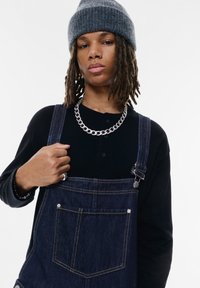 Salopette in denim con bretelle regolabili, di colore blu scuro, tasca frontale, indossata sopra una maglietta nera a maniche lunghe, collana in argento, berretto di maglia grigio.