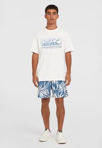 Weisses Baumwoll-T-Shirt mit blauem grafischen Druck; kombiniert mit blauen Badehosen, die ein weisses tropisches Blattmuster aufweisen; weisse Sneakers.