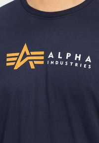 Μπλε βαμβακερό μπλουζάκι με μεγάλο πορτοκαλί λογότυπο που περιλαμβάνει έναν αφηρημένο "Α" και ριχτές λεπτομέρειες, μαζί με το κείμενο "ALPHA INDUSTRIES" σε λευκό χρώμα.