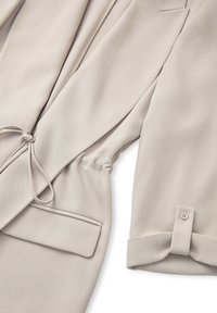 Beige Jacke aus strukturiertem Stoff, mit Kordelzug in der Taille und umgeschlagenen Manschetten. Merkmale sind eine Seitentasche und dezente Nahtdetails.