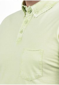 Lichtgroene polo met een knoopsluiting, een voorzak en een gestructureerde stof. Bevat vier knopen en heeft een licht vervaagde uitstraling.