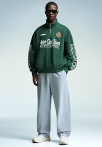 Mand iført en mørkegrøn half-zip sweatshirt med hvid tekst og grå brede sweatpants, hvide sneakers og sorte solbriller, står selvsikkert.
