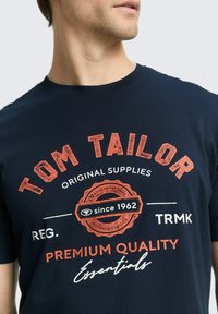 Granatowy bawełniany t-shirt z wyrazistymi pomarańczowymi i białymi grafikami oraz tekstem: "TOM TAILOR", "PREMIUM QUALITY" i "since 1962". Okrągły dekolt.
