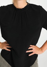 Personne portant un haut texturé à manches courtes noir avec un col froncé, les mains sur les hanches, et un pantalon beige devant un fond uni.