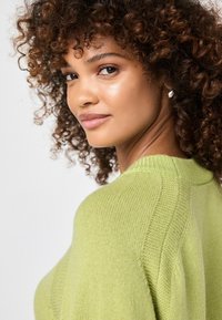 Junge Frau mit lockigem Haar, die einen hellgrünen Strickpullover trägt, schaut über ihre Schulter mit einem leichten Lächeln vor einem einfarbigen Hintergrund.
