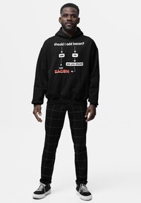 Uomo che indossa un hoodie nero con un diagramma di flusso sul bacon che dice "dovrei aggiungere bacon?" e pantaloni neri con righe bianche a griglia, in piedi su uno sfondo bianco.