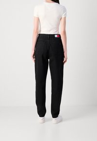 Femme portant un jean noir taille haute avec poches arrière et un t-shirt blanc à manches courtes, debout devant un fond blanc uni.