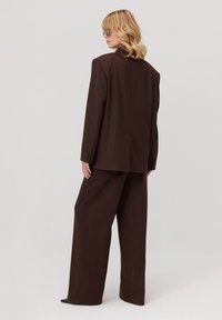 Kobieta z blond włosami, ubrana w ciemno-brązowy oversize'owy garnitur i okulary przeciwsłoneczne, stoi tyłem na jednolitym jasnym tle.