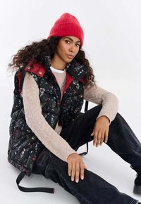 Czarna pikowana kamizelka w kwiaty z czerwoną podszewką i wysokim kołnierzem, zestawiona z jasnobeżowym swetrem w wyplatany wzór i ciemnymi dżinsami. Na głowie czerwona czapka zimowa.