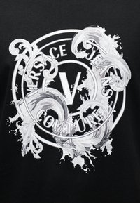 Λευκά κύματα με περιστρεφόμενα διακοσμητικά μοτίβα περιβάλλουν ένα κυκλικό λογότυπο Versace Jeans Couture με ένα κεντρικό γράμμα V σε μαύρο φόντο.