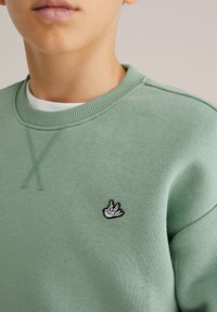 Zelená mikina z mäkkej tkaniny s rebrovaným výstrihom a malým vyšívaným logom vtáčika na hrudi.