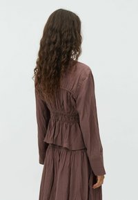 Long-sleeve, brown textured top with gathered waist and peplum detail. Features soft fabric and a relaxed fit, seen from the back.