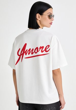 Donna con taglio di capelli a caschetto nero che indossa occhiali da sole rotondi e una maglietta bianca con "Amore" in scritta rossa sulla schiena.
