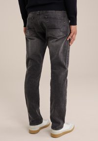 En person i mörkgrå jeans och vita sneakers står med ryggen mot kameran mot en beige bakgrund.