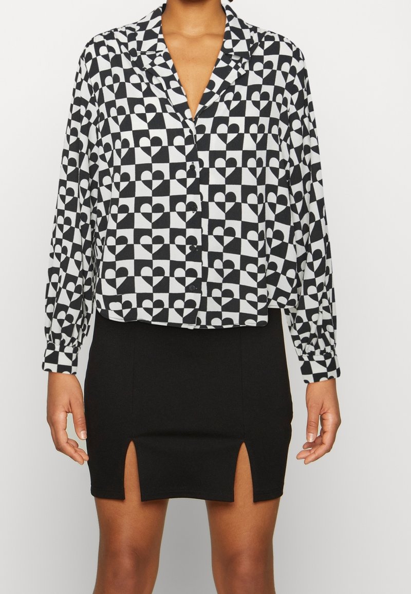 Chemise boutonnée noir et blanc avec un motif de cœur géométrique et des manches longues. Associée à une jupe noire ajustée avec des fentes sur les côtés.