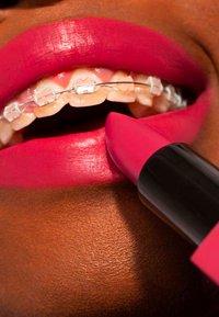 Jasnoróżowa szminka nakładana na błyszczące usta z aparatem ortodontycznym; skóra ma gładką, ciepłą teksturę i subtelny połysk.