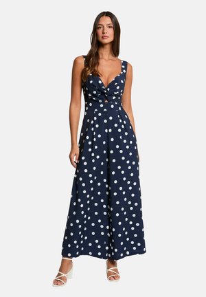 Donna che indossa una tuta blu navy a gamba larga con pois bianchi, un'apertura frontale annodata e sandali bianchi con tacco.