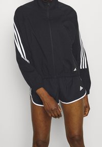 Schwarze Sportjacke mit hohem Kragen, Reißverschluss und weißen drei Streifen an den Ärmeln. Passende schwarze Shorts mit weißem Saum.