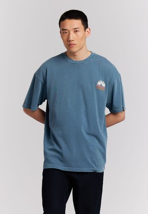 Jonge man staat met handen achter de rug, draagt een blauw oversized T-shirt met een klein logo op de borst en donkere broek.