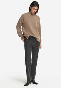 Beiger Strickpullover mit hohem Kragen, lockerer Passform und gerippten Bündchen, kombiniert mit dunkelgrauen, gerade geschnittenen Jeans und schwarzen, spitzen High Heels.