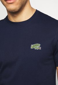 Mörkblå bomullströja med rund halsringning, prydd med en grön och vit broderad "Lacoste"-logotyp med en krokodildesign på bröstet.