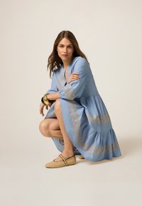 Vestido azul claro con bordados geométricos dorados, mangas largas y falda en capas. Zapatos planos de mimbre beige con un acento de hebilla.