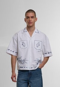 Witte short-sleeve shirt met blauwe borduursels, voorzien van twee borstzakken, bloemenpatronen op de mouwen, en een olijftakontwerp aan de zoom.