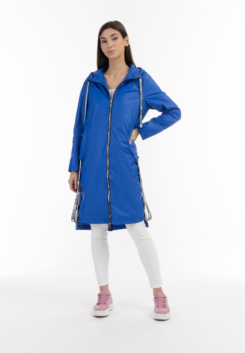 Blauwe waterdichte lange jas met een rits, capuchon en trekkoorden, met zijzakken en contrasterende witte hardwaredetails. Draagt over witte leggings en roze sneakers.