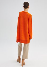 Przewymiarowany pomarańczowy sweter dziergany o fakturze prążkowanej, z okrągłym dekoltem i długimi rękawami, zestawiony z jasnobeżowymi spodniami i butami na obcasie.