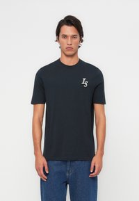 Black cotton t-shirt with a crew neck, short sleeves, and a small embroidered logo on the left chest, paired with blue denim jeans.