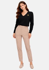 Femme aux longs cheveux ondulés portant un pull noir à col en V avec un détail soleil doré, un pantalon beige sur mesure et des mocassins beige, debout avec assurance.