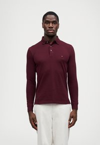 Camisa pólo de manga longa em algodão bordeaux com colarinho, botões de três e pequeno detalhe com logotipo no peito. Combinada com calças de cor clara.