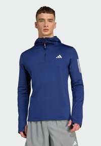 Tamnoplavi sportski pulover s djelomičnim zatvaračem, kapuljačom i teksturiranim materijalom; sadrži bijeli Adidas logo i tri pruge na lijevom rukavu.