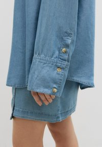 Helles blaues Denim-Hemd mit lockerer Passform, langen Ärmeln mit drei Knopfmanschetten und geradem Saum. Der Stoff hat eine weiche Textur.