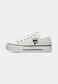 Witte lage sneaker met dikke zool, zwarte strepen detail en kleine cartoonfiguur met zonnebril aan de zijkant.