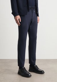 Σκούρο μπλε παντελόνι με ρυθμιζόμενη μέση, κομψή γραμμή και πλάγιες τσέπες, συνδυασμένο με μαύρα δερμάτινα παπούτσια με λεία υφή.