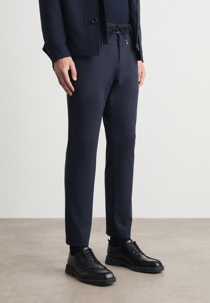 Σκούρο μπλε παντελόνι με ρυθμιζόμενη μέση, κομψή γραμμή και πλάγιες τσέπες, συνδυασμένο μ�ε μαύρα δερμάτινα παπούτσια με λεία υφή.