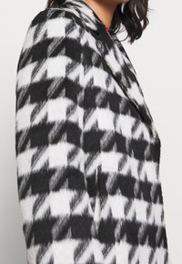 Sort-hvid houndstooth-mønstrede uldfrakke med skrå krave, dobbelte knapper og ensartet stofstruktur. Synlig sidelomme.