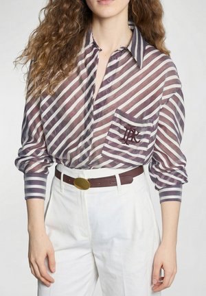 Femme portant une chemise à rayures diagonales marron et blanche boutonnée avec une poche devant, et un pantalon blanc taille haute avec une ceinture marron.