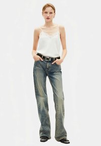 Bijeli top s čipkastim obrubom u stilu kame, uparen s izblijedjelim plavim trapericama ravnih nogavica, upotpunjen crnim remenom i crnim gležnjačama.