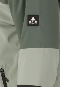 Groene waterdichte jas met een opvallend zwart "Whistler" logo patch, ritszakdetail en contrasterende lichtere mouwsecties.
