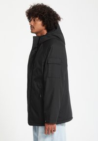 Homme aux cheveux bouclés portant une veste à capuche noire avec une poche sur la manche, debout de profil sur un fond blanc.