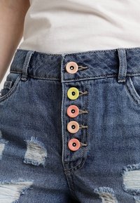 Shorts in denim con are invecchiato, con cinque bottoni colorati rosa, giallo e verde sul davanti. Design classico a cinque tasche.