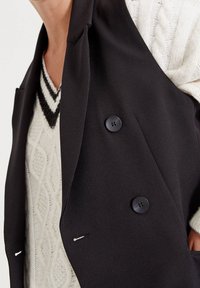 Czarna marynarka z podwójnymi guzikami i ostrymi klapami, w połączeniu z kremowym swetrem w warkocze z dekoltem w kształcie V i kontrastowymi czarnymi paskami.