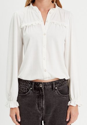 Femme portant un chemisier blanc à manches longues boutonné avec des poignets volantés et un jean noir taille haute.