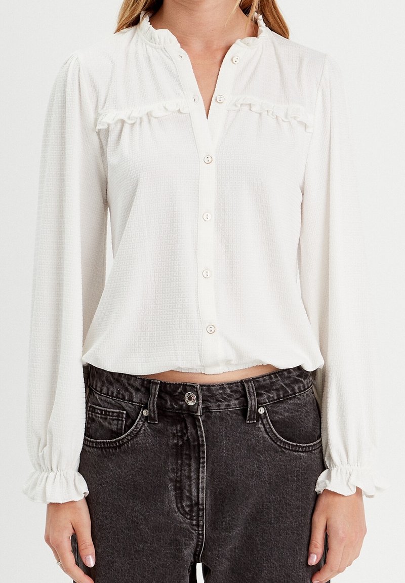 Femme portant un chemisier blanc à manches longues boutonné avec des poignets volantés et un jean noir taille haute.