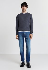 Mann, der einen schiefergrauen Pullover, ein weißes Unterhemd, blaue Jeans und schwarze Stiefel trägt, steht vor einem einfachen hellen Hintergrund.